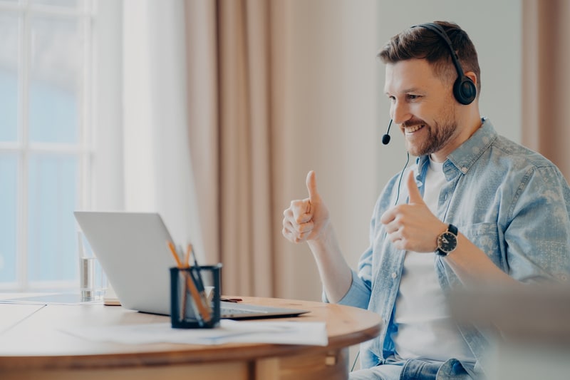 Satisfied bearded male model attends online training from home uses headset makes like gesture with both hands looks attentively at laptop computer makes video call poses against cozy interior