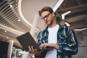 Bottom view of caucasian guy watching something on laptop computer in office space. Concept of flexible workplace. Idea of freelance and remote work. Young focused bearded successful man in glasses