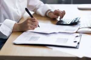 Close-up photo. Beautiful hands of a young woman in a white shirt. They write documents with a pen, calculate on a calculator at the table in the office, where there is a computer, a cup of coffee.