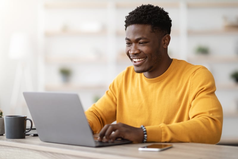 Successful black guy project manager sitting at workdesk at office, typing on laptop keyboard and smiling, chatting with clients, enjoying his job, copy space. Freelancer working from home