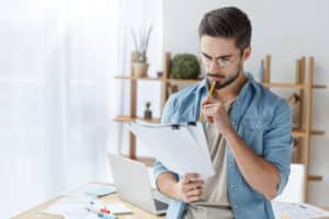 Young male executive officer has attentive look at documents, reads details of agreement, studies contract with focused expression, holds pencil. Fashionable man works on creating annual report