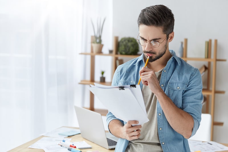 A man in glasses is holding a clipboard and looking at project management software.