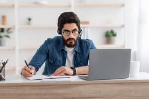 Millennial arab guy in headset watching online webinar on laptop at home, having online training, studying with computer and taking notes to notepad, copy space