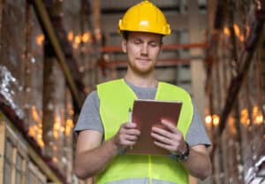 Man looking at check sheet to checking inventory by lablet. A young caucasian man wearing helmet and reflective jacket working in a warehouse.