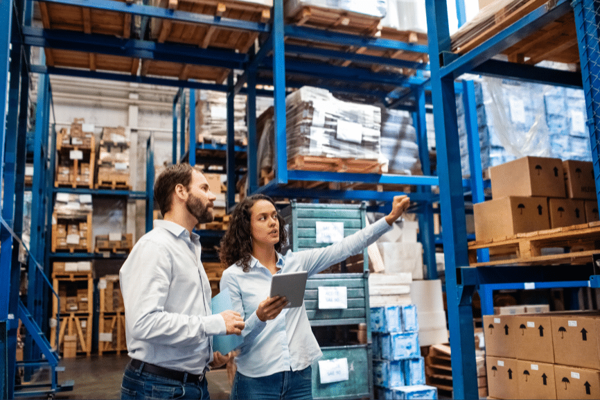 Two warehouse workers, a man and a woman, are inspecting inventory on blue metal shelving. The woman holds a tablet and points towards the shelves, while the man looks in the same direction. Stacks of boxes and various packed items fill the shelves around them as they reshape your inventory and supplies management.
