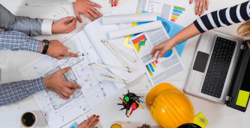 A group of people collaborating over a table filled with architectural blueprints, a laptop, vibrant charts, and a yellow hard hat. With coffee cups in hand and plans unfolding before them, hands are actively pointing and discussing how to ebase projects. A small plant adds a touch of green to the action-packed scene.