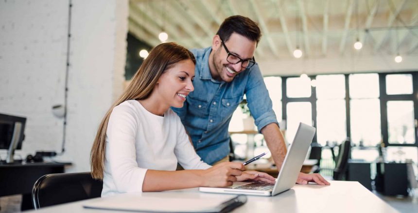 A woman and a man are smiling while working together at a desk with a laptop in a modern office. The woman is typing, and the man stands next to her holding a pen, both appearing engaged and collaborative, perhaps brainstorming ideas for smarter scheduling in the New Year.