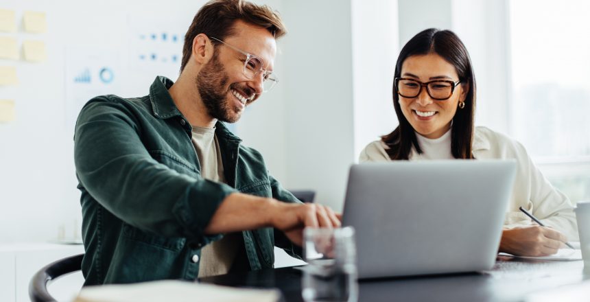 Two business people using a laptop together while sitting in a meeting. Happy business people looking at a slide presentation in an office.