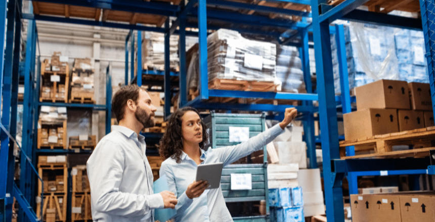 Two warehouse workers, a man and a woman, are inspecting inventory on blue metal shelving. The woman holds a tablet and points towards the shelves, while the man looks in the same direction. Stacks of boxes and various packed items fill the shelves around them as they reshape your inventory and supplies management.