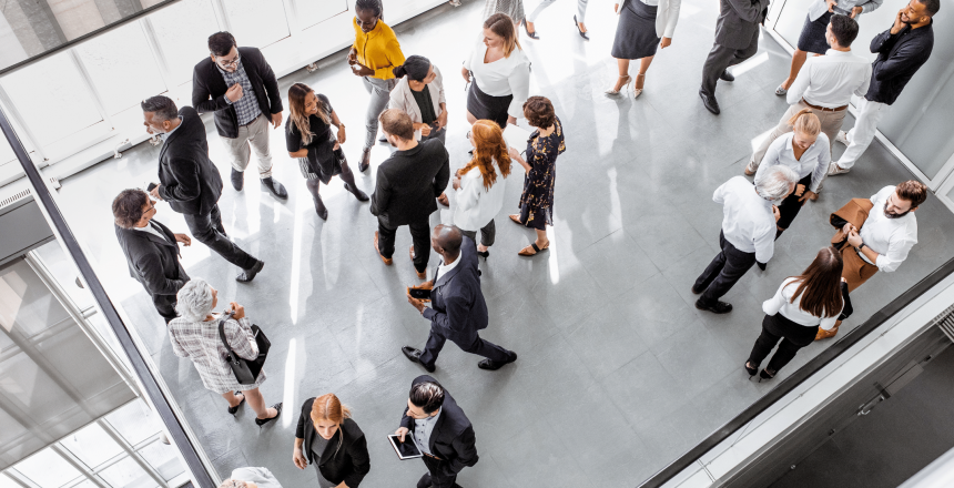 An overhead view of a modern office space bustling with activity during the ebase Spring Tradeshow Season 2024. Many people are mingling—some talking in groups, while others walk through the area. The gray floor and natural light from large windows accentuate the diverse mix of individuals.