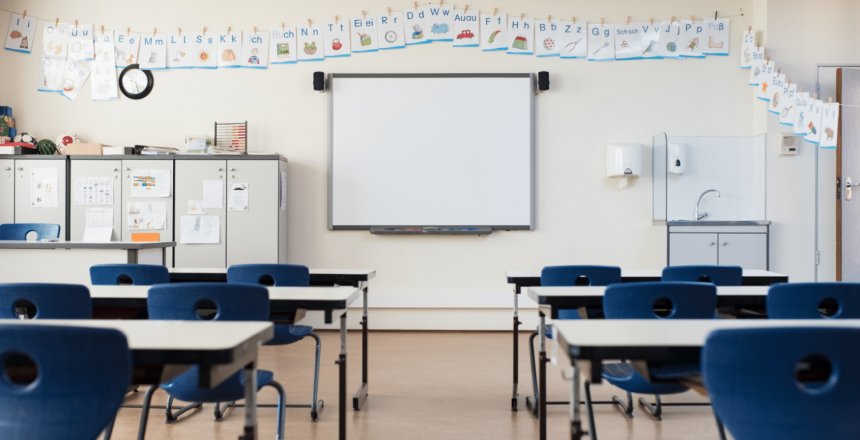 A classroom with a whiteboard at the front, surrounded by alphabet posters along the walls. Blue chairs and white desks are arranged in rows. Cabinets and a clock are on the left side, with a sink and sanitizer dispenser on the right wall.